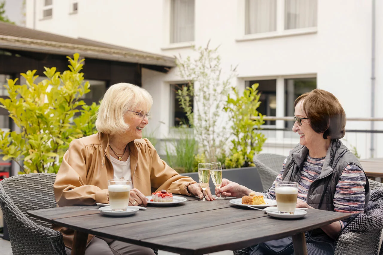 Zwei Seniorinnen trinken fröhlich Kaffee auf der Terrasse des Café Sophie in der der Alerds-Stifung Bezahlbare Pflege mit einem neuen Konzept